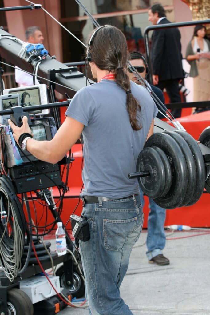 Dos d'opérateur de grue caméra avec écouteurs, ponytail, mains sur les commandes. Poids (5KG) et écrans visibles sur l'équipement.