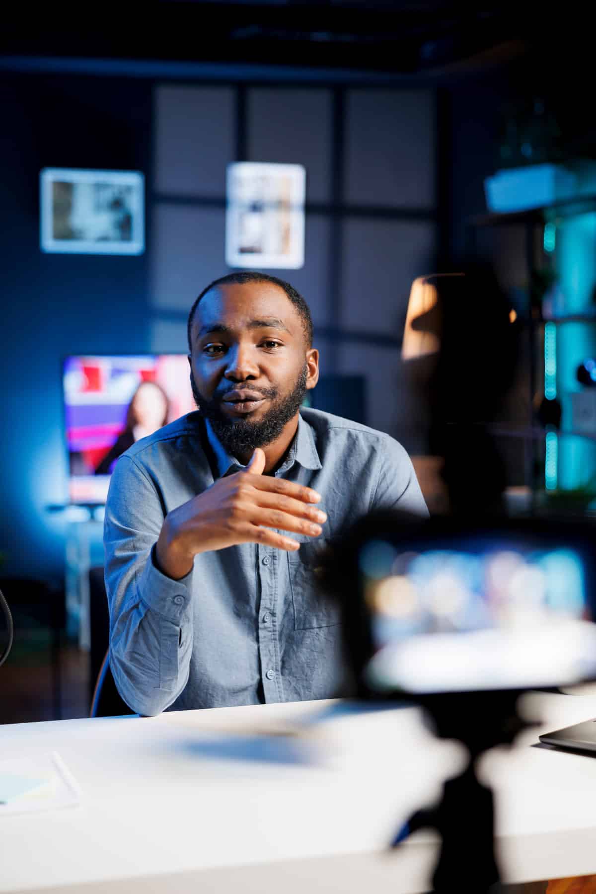 Homme en chemise bleue parlant et gestuant devant une caméra smartphone. Studio sombre avec éclairage bleu et écran en fond.