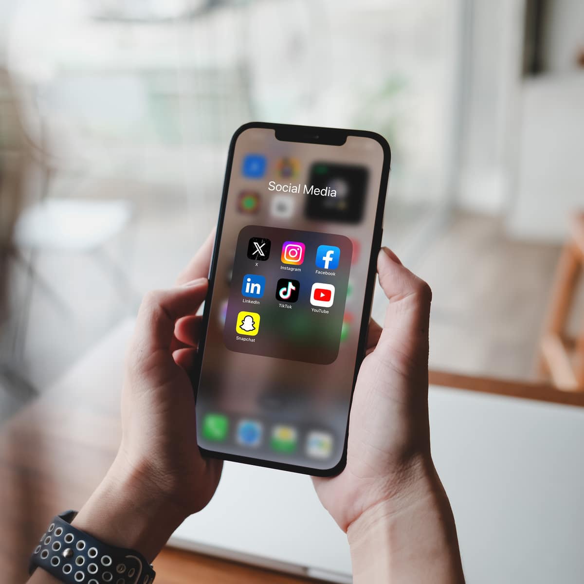 Personne tenant un téléphone affichant un dossier 'Social Media' avec les icônes X, Instagram, Facebook, LinkedIn, TikTok, YouTube, Snapchat.