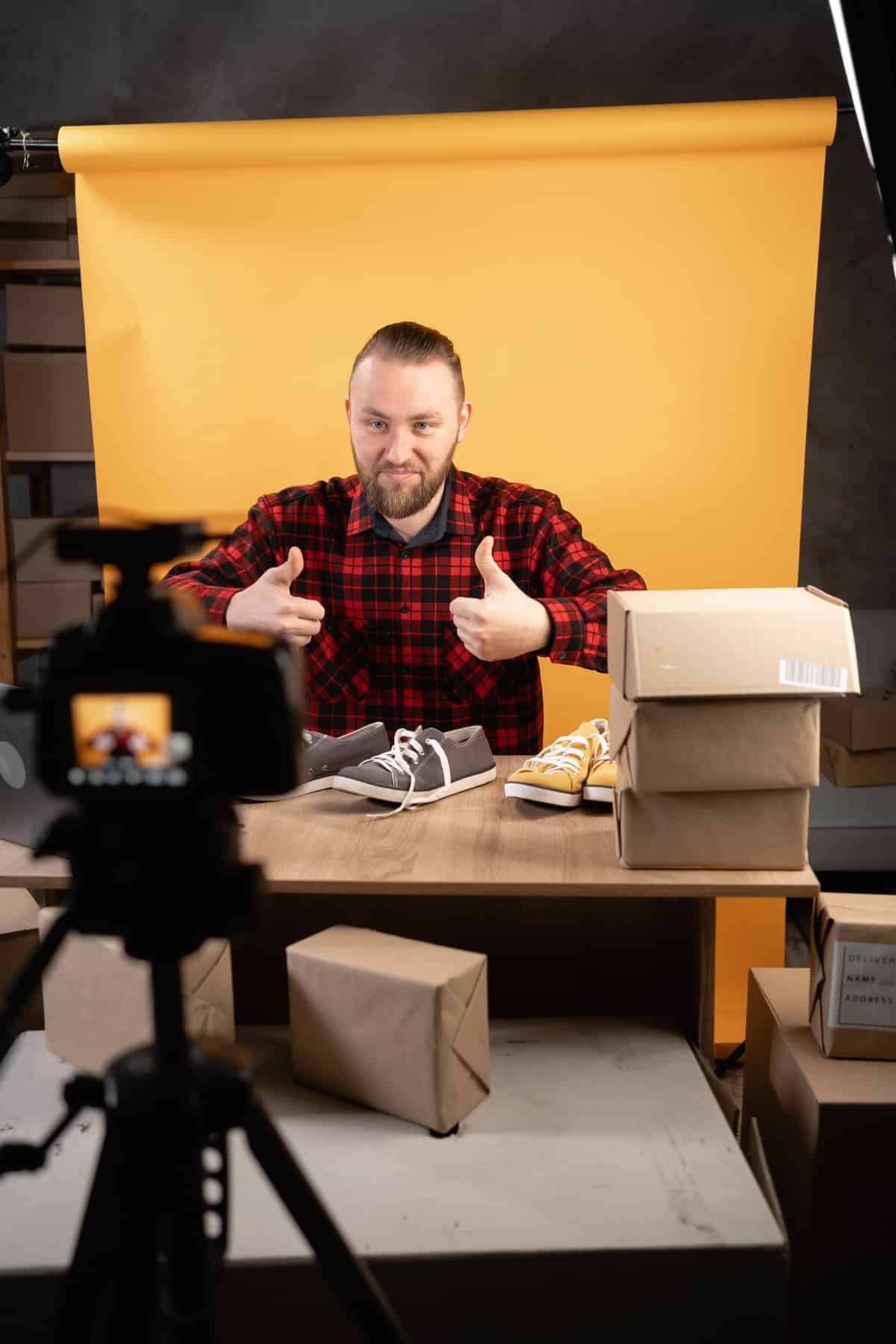 Homme en chemise à carreaux pouce levé, filmant une vidéo d'unboxing/e-commerce avec chaussures et cartons d'expédition sur un bureau.