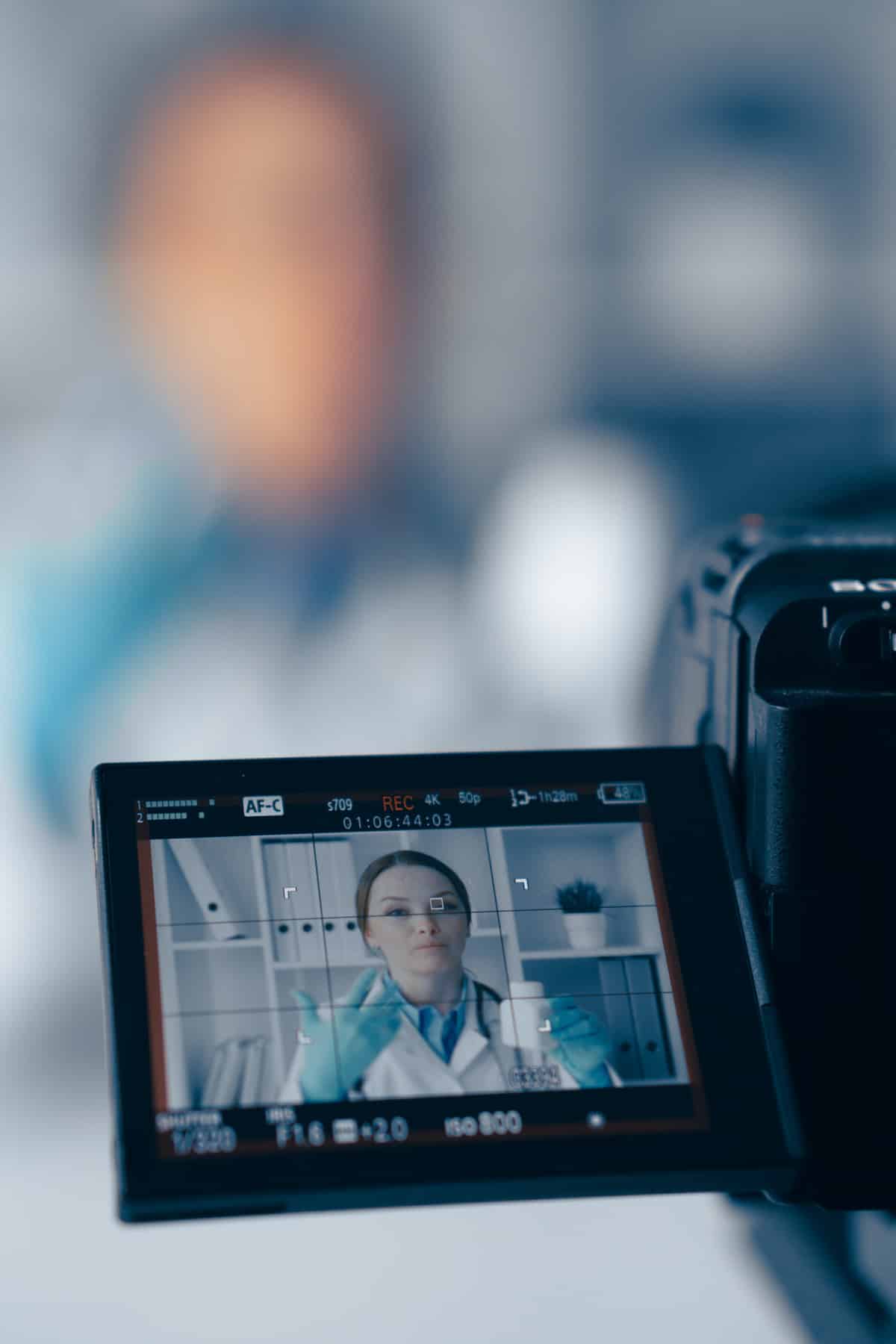 Écran caméra REC: médecin femme en blouse, gants bleus, expliquant avec bouteille blanche. Cadran de mise au point visible.